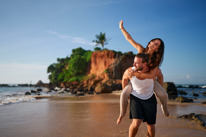 Joyful couple enjoys piggyback ride on tropical beach, woman raises hand high, man carries partner by sea. Romantic getaway, playful mood, sandy shore, love adventure. Tourists explore island paradise